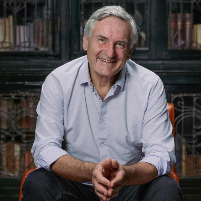 Photograph showing an older gray-haired man seated with hands together and smiling at camera. He wears a light blue button-down shirt and dark pants, with enclosed glass bookcases in the background.