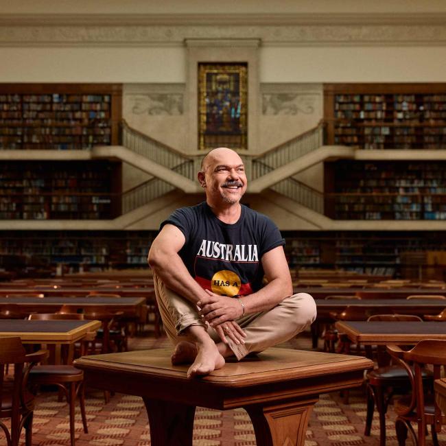 Photograph of a smiling bald man seated cross-legged atop a wooden table inside an ornate library filled with books and balconies. He wears beige pants with a black t-shirt featuring the Aboriginal flag and is looking to the side.