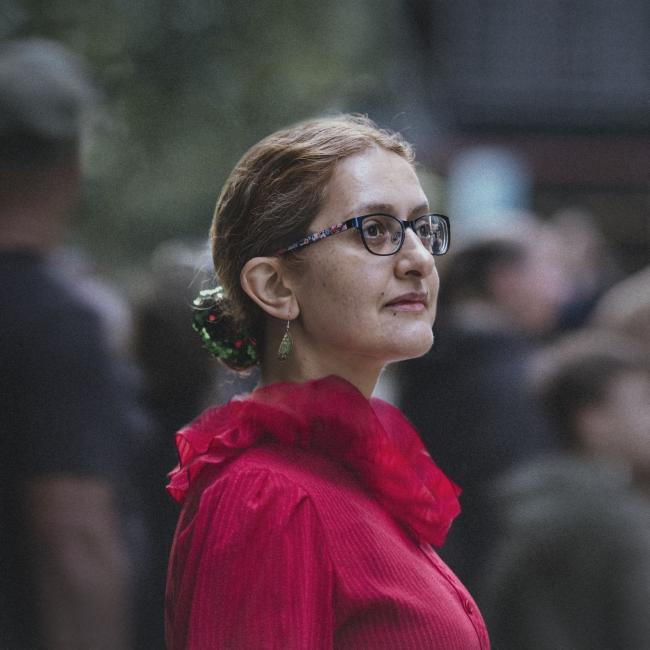 Photograph of a woman wearing glasses and a bright red blouse with ruffled collar standing outdoors among a blurred crowd. Focus is on the upper body and profile, highlighting a green hair accessory and dangling earrings against a soft, natural background.