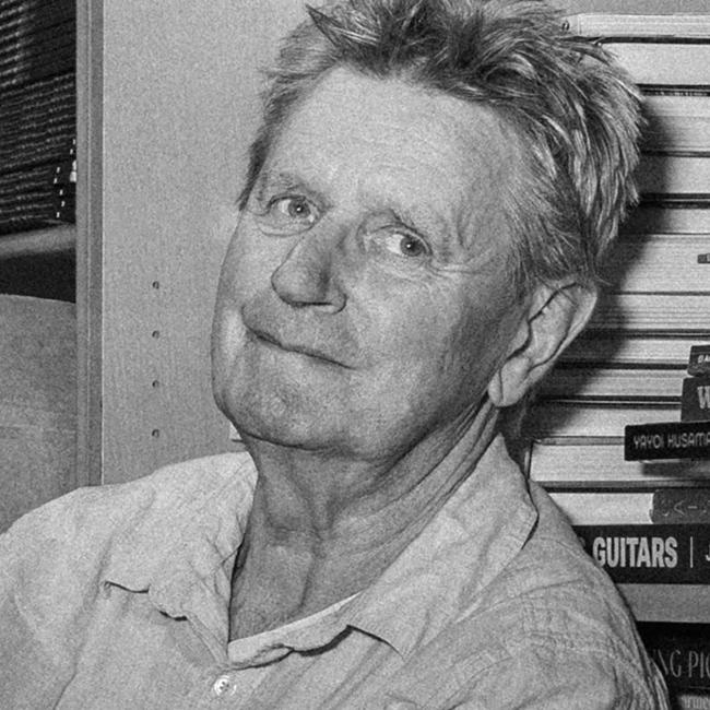 Photograph of a man sitting on a box in a cluttered storage room filled with shelves of books and stacked cardboard boxes. The person is casually posed with arms crossed, surrounded by various items including a trolley and scattered books on the floor, suggesting a setting related to book storage or inventory.