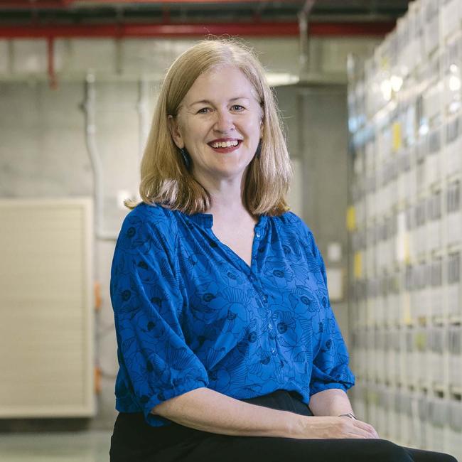 Photograph of a woman sitting on a red stool in a large archive room filled with tall shelves of organized white boxes. The setting highlights a structured storage system with labeled boxes and fluorescent lighting, emphasizing an environment focused on document preservation and organisation.