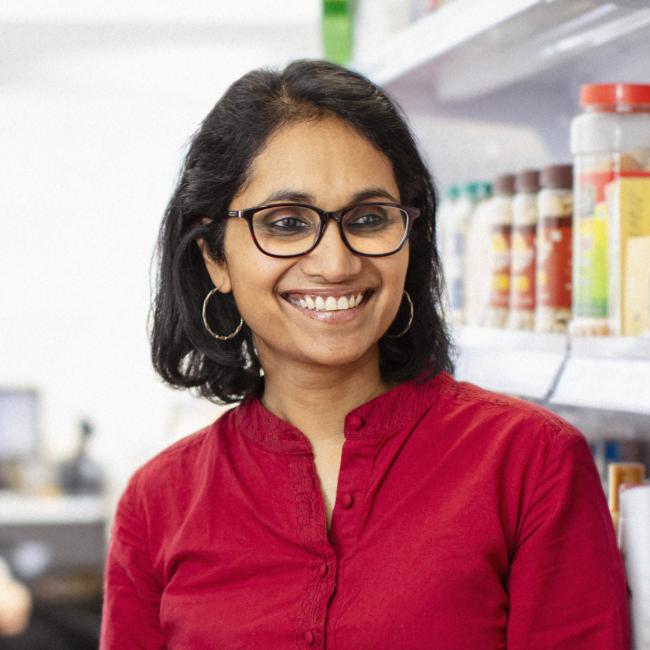 Photograph of a woman wearing a red shirt and glasses standing next to shelves stocked with various packaged products. Shelves contain colorful boxes and bottles arranged in rows, indicating a retail environment focused on health or beauty item