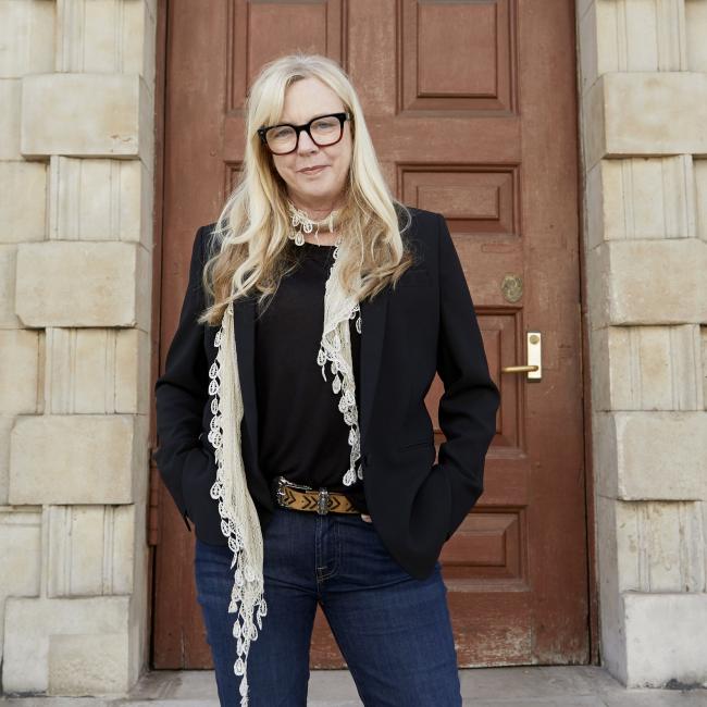 Photograph of a woman standing confidently on stone steps in front of a large wooden door set in a sandstone building with classical architectural details. The person wears a black jacket, dark jeans, red ankle boots, and a light scarf, positioned centrally under a large hanging lamplight. 